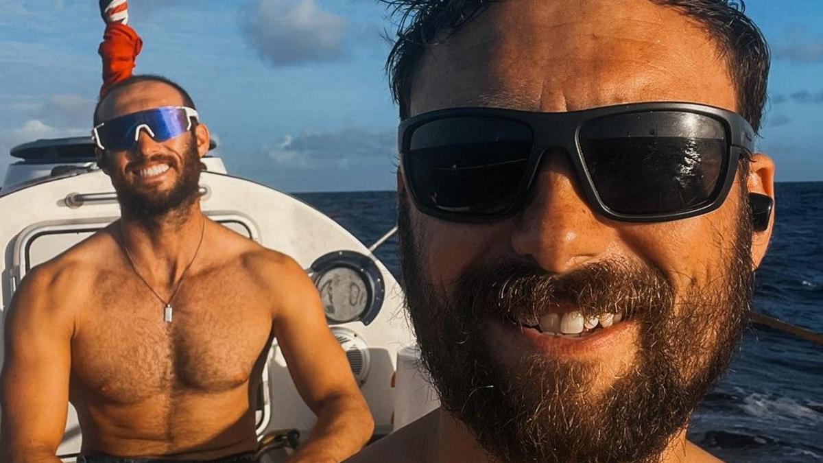 Two men with beards and wearing sunglasses are smiling at the camera. Mr Mason is in the foreground and on the photo a pole, containing a flag, is directly above and behind Mr Mattock's head. The sea is behind the rowing boat they are in.