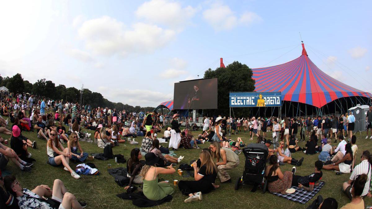 Concert goers sitting on the grass drinking alcohol and watching an artists perform on stage via a screen. There is a red and blue tent in the background.