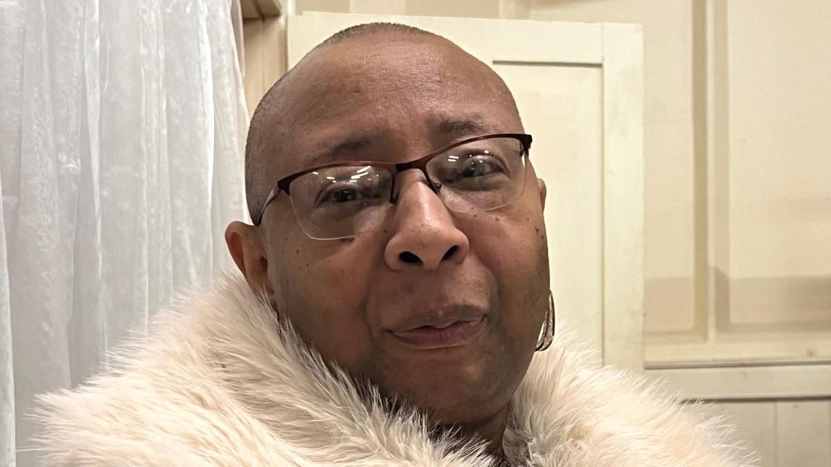 Marsha wearing a cream faur jacket and black glasses as she looks at the camera with a white wall and cream door behind her