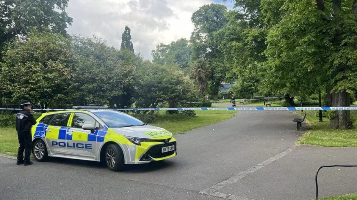 A police officer stands by a marked police car guarding a taped section of East Park