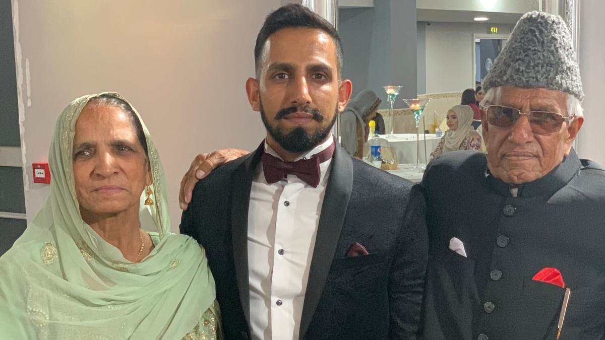 A family picture of a young man with two older people, a man and a woman, either side of him. They are at some sort of celebration. The young man wears a tuxedo and bow tie. The older couple wear traditional South Asian formal clothes, the man in a buttoned jacket and conical cap, the woman a green South Asian outfit with gold embroidery. Behind them are other people in glamorous outfits.