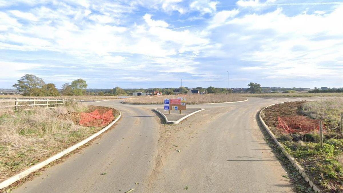 A road with grass verges either side and a roundabout is in the distance.