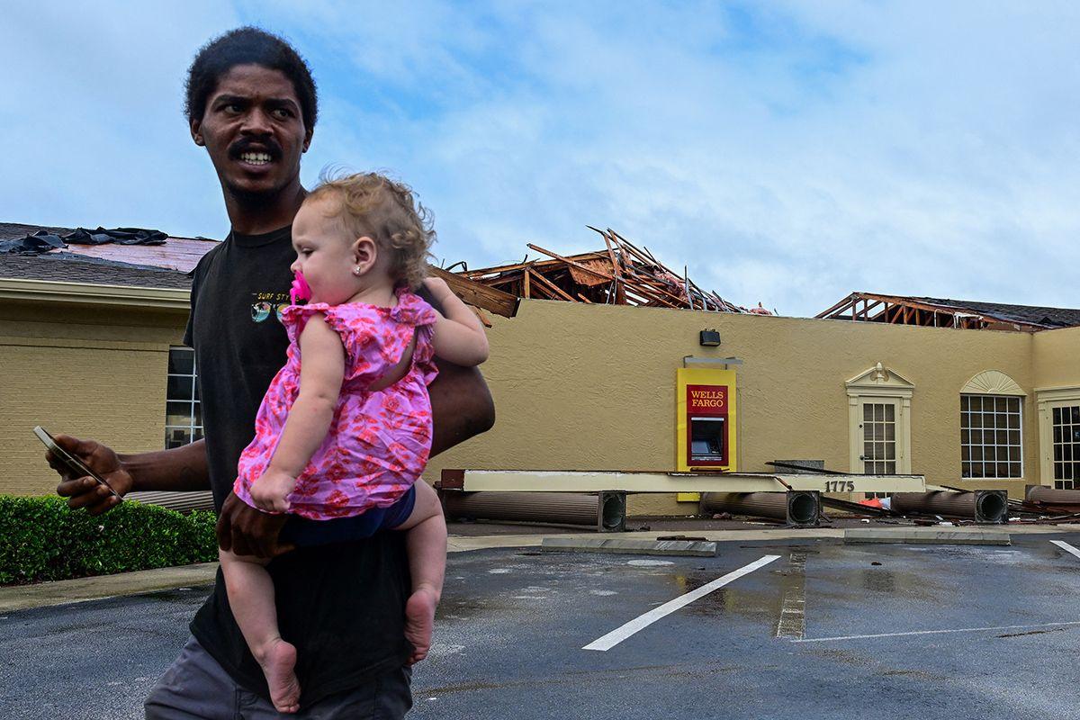 A man walks while carrying a baby with a Wells Fargo bank branch in the background with its roof in splinters