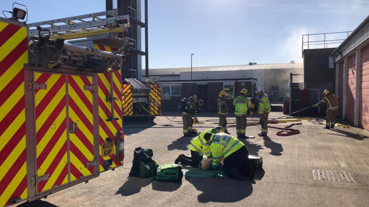 Several fire fighters and paramedics carrying out a demonstration in the yard of a fire station, with a dummy lying on the ground receiving CPR. There is a fire engine just visible in the foreground on the left, and behind it, another engine and the base of a fire training tower