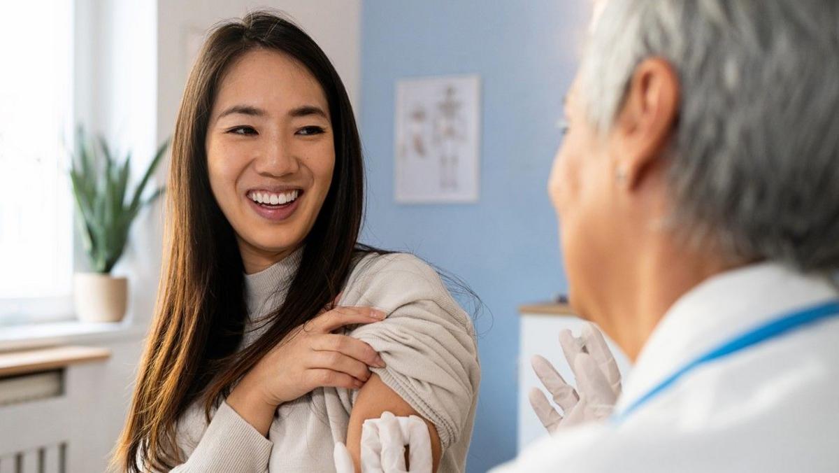 A woman smiles as she holds up her sleeve to be given a flue vaccine by a health worker who is wearing gloves, a white coat and a lanyard. They are in a consulting room with a plant on the windowsill.