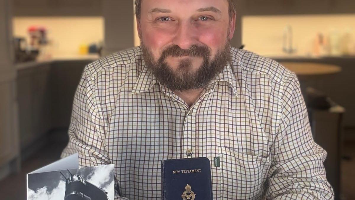 Ross McEwen poses with the bible and a picture of a Shorts Sterling. He has a beard and wears a brown and white checked shirt.