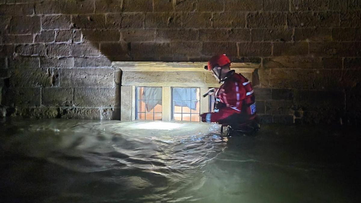 A member of Northamptonshire Search and Rescue wades through flood water. He is wearing red high-visibility clothing and a red hard hat. He is holding a torch while walking past a window, where the flood water is high enough to have covered part of it.