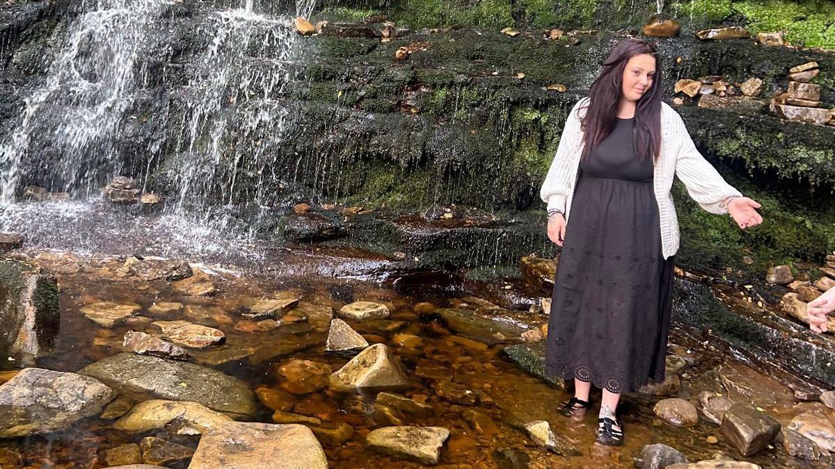 Bethany Wooldridge in the outdoors. She is stood next to a waterfall and has one hand outstretched. She is wearing a long black dress and a cream cardigan, and has long brown hair. 