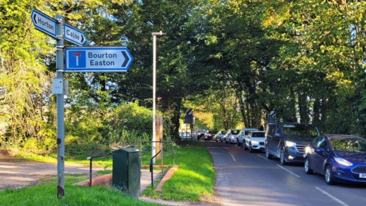 Queues of traffic entering Bishops Cannings amid the diversion with trees and grass on either side of the road. A sign points in the opposite direction to Calne while two other signs point to Horton and to Bourton and Easton.