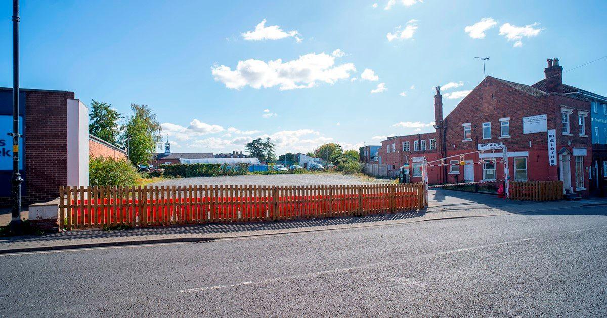 A flat gravel site behind a wooden fence adjacent to a road, with an entrance barrier shut to cars. A red-brick two-storey building to the right has a sign for Bridge Street pay and display half torn away. Greenery and other buildings can be see in the background.