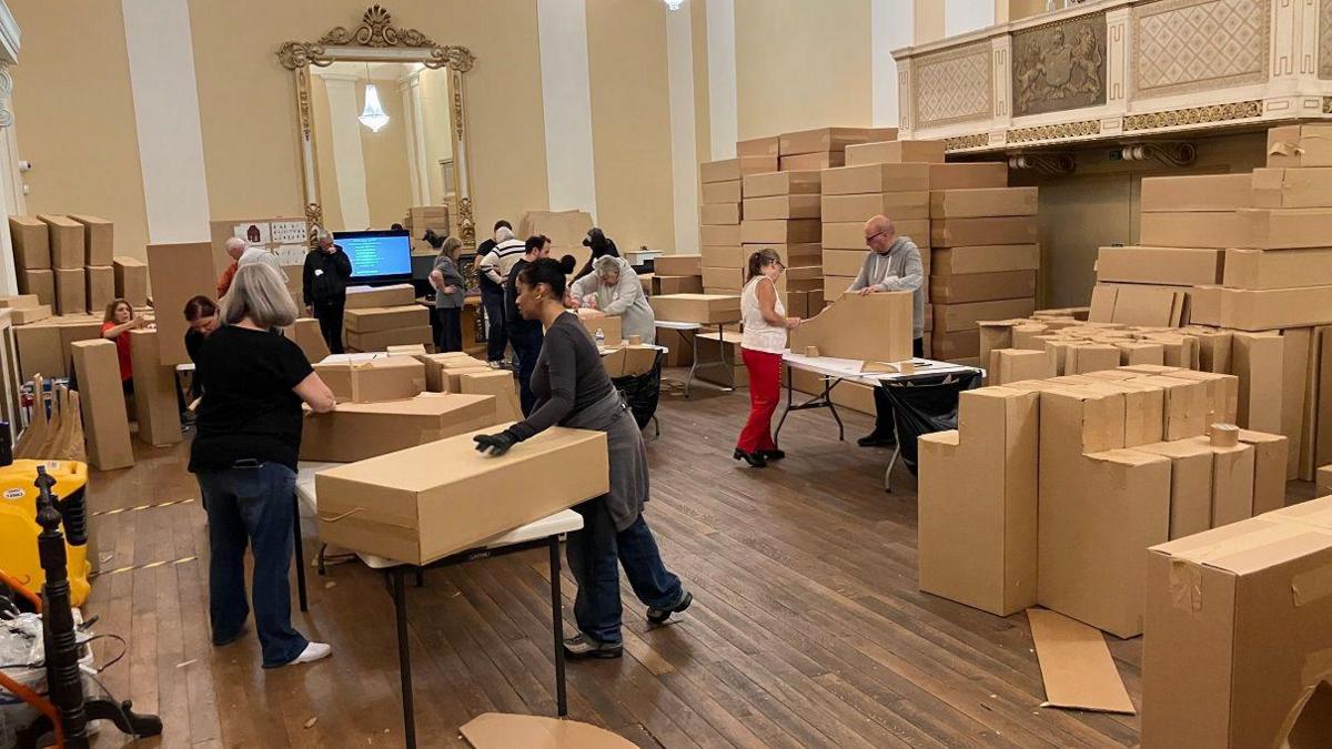 People are using cardboard to create various structures which will be part of the finished work. Many have gloves and they are working in pairs. The large room is full of stacked-up boxes.