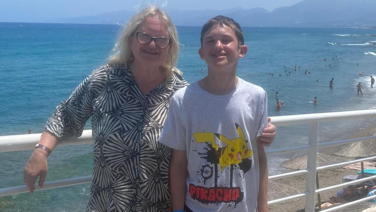 A woman poses for a photo with her arm around her son. Both are smiling, with a beach and swimmers in the background. The weather looks warm, with both wearing summer clothing.