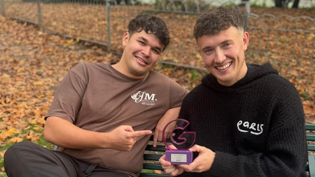 A man in a brown T-shirt and short black curly hair points at another man holding a purple award that is shaped like the letter 'G'. He is wearing a black hoodie and brown has short brown curly hair.