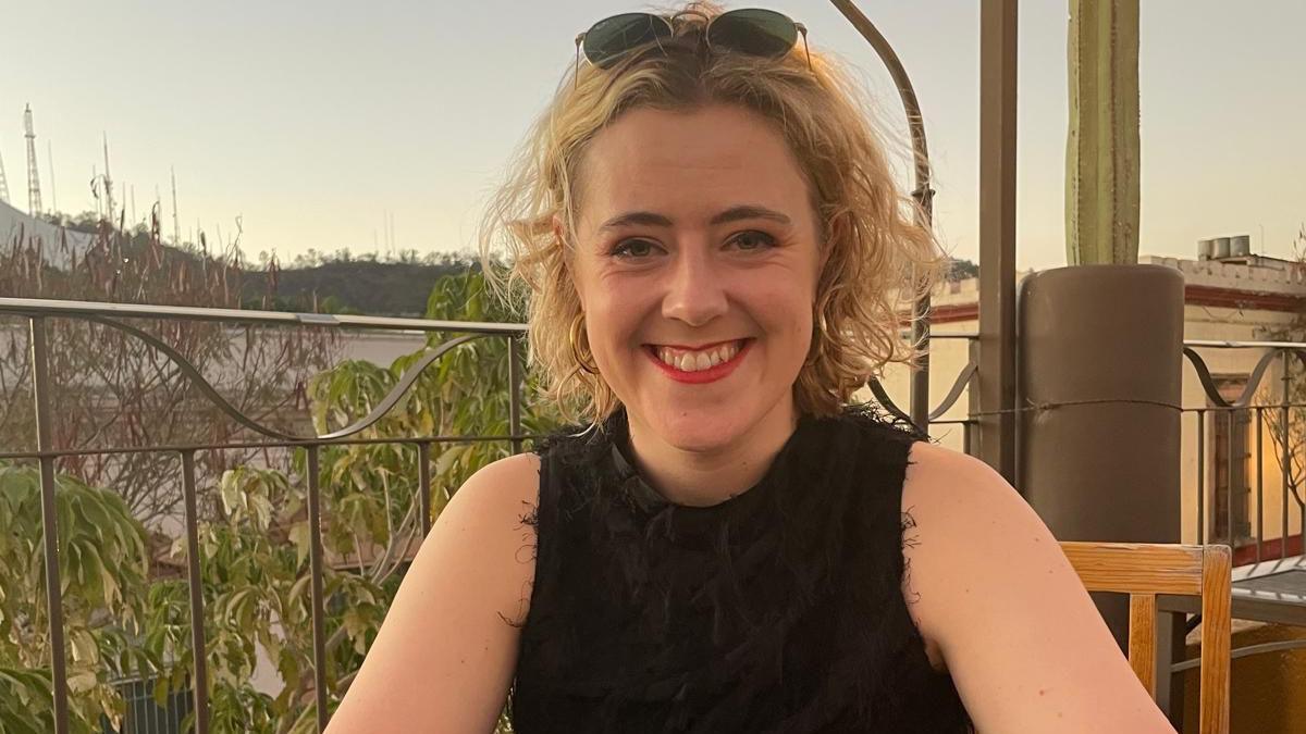 Meg has short blonde hair and has sunglasses sat on top of her heard while smiling at the camera. She is sat at a restaurant table outside.