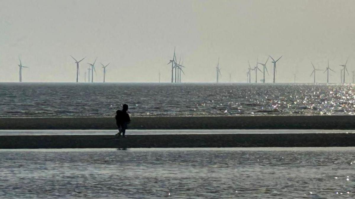 A person in silhouette walks along a sand bank on Crosby beach with water pooling on either side and the sea, with an off shore wind farm, in the distance