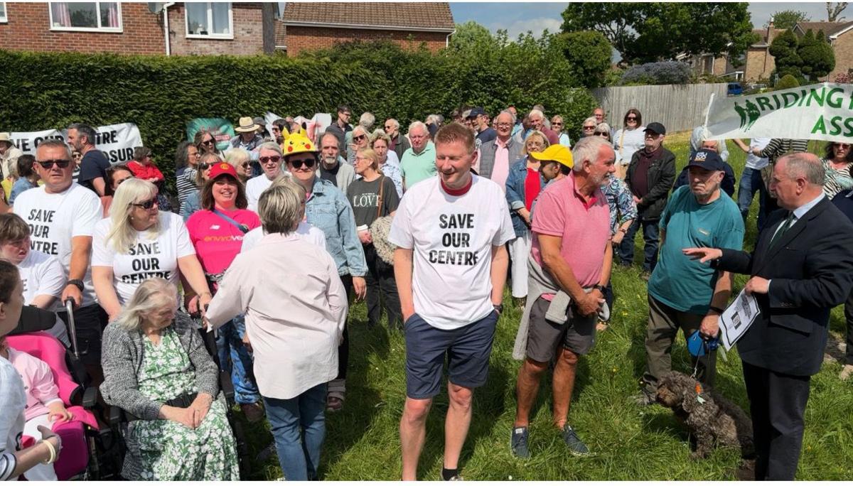 A large group of supporters of the Blandford Community Centre, some wearing T shirts with "Save Our Centre" on them. Local MP Simon Hoare is seen front-right talking to protestors