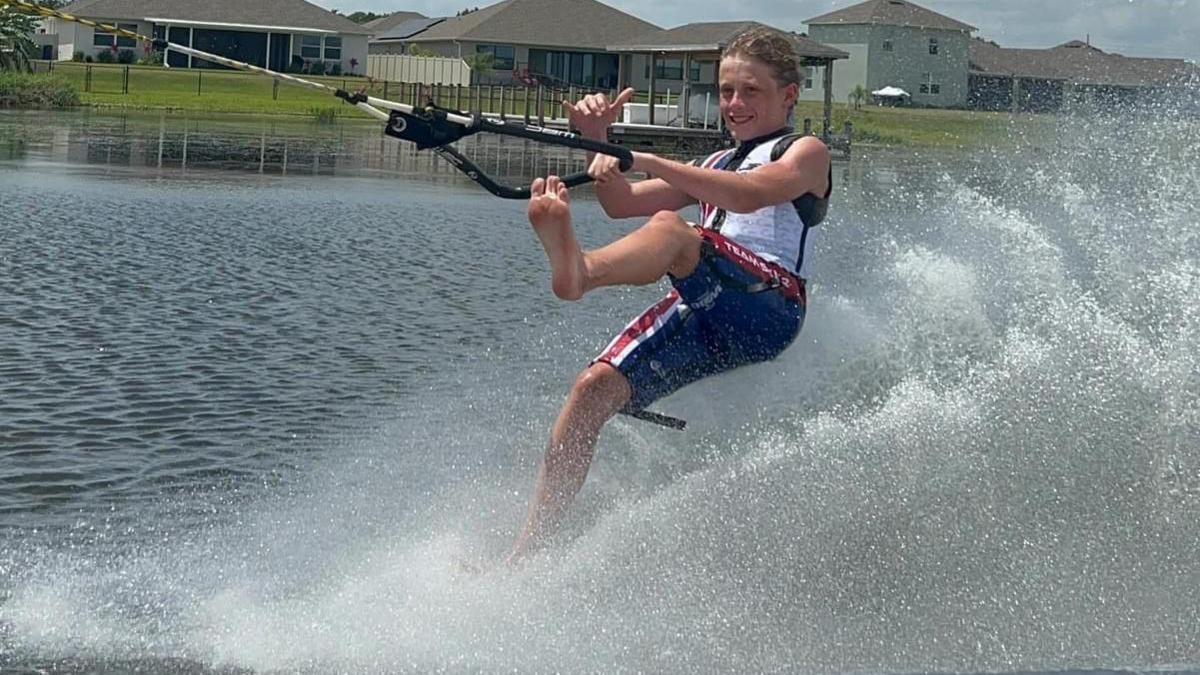 Olly Moore is pictured in a white and blue suit holding onto a rope on the back of a boat and he skis using just his feet. He has one foot up in the air as water sprays behind him.