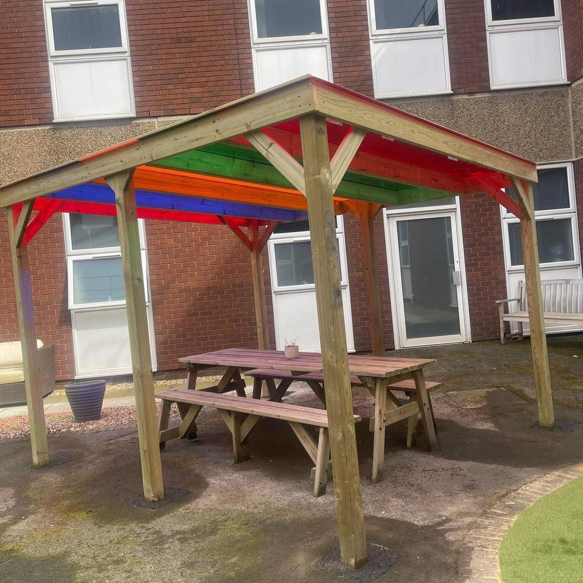 A wooden shelter with a roof painted red, green, orange and blue in front of a building with multiple windows. Underneath the shelter is a wooden bench.