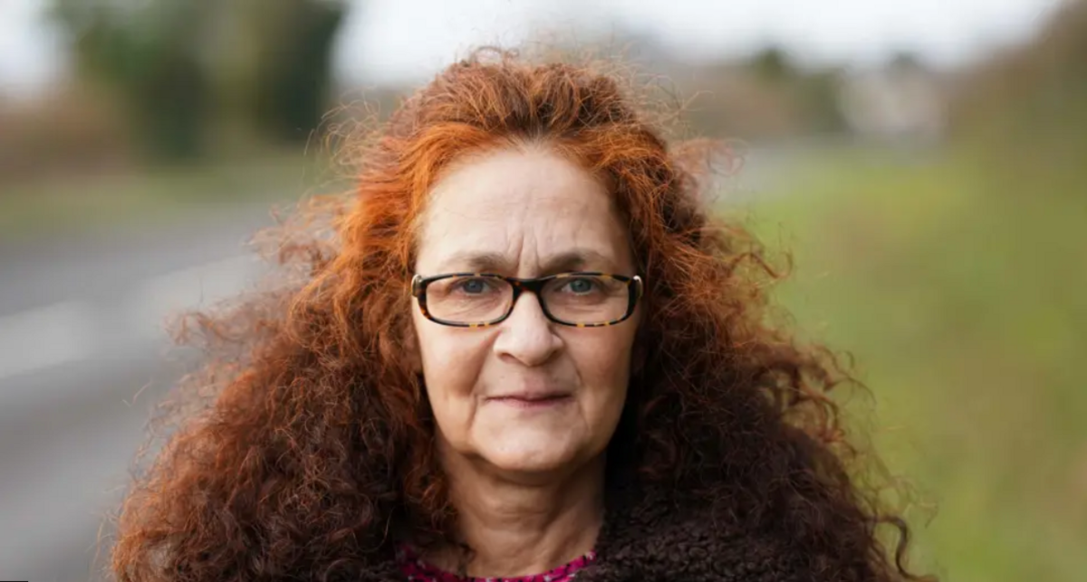 Woman with rectangle frame glasses and thick curly red hair. she is wearing a brown teddy bear fur coat with a pink shirt underneath