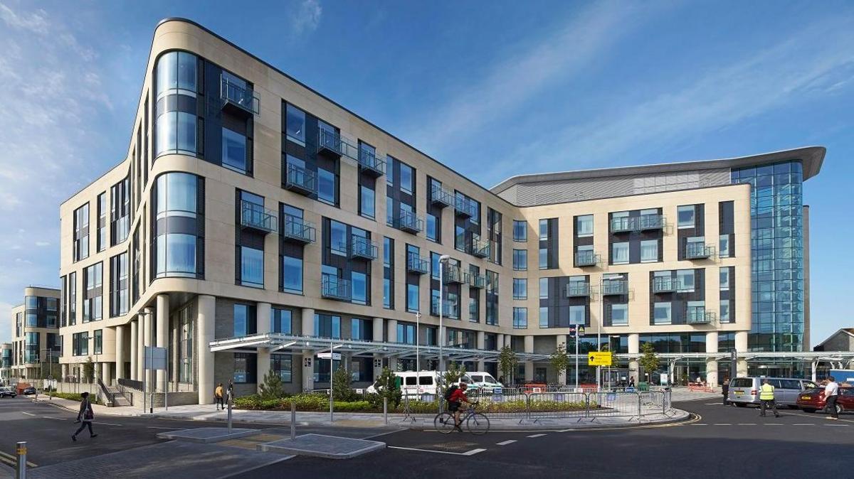 Southmead Hospital in Bristol which has multiple stories with glass windows and cars and vans parked outside. 