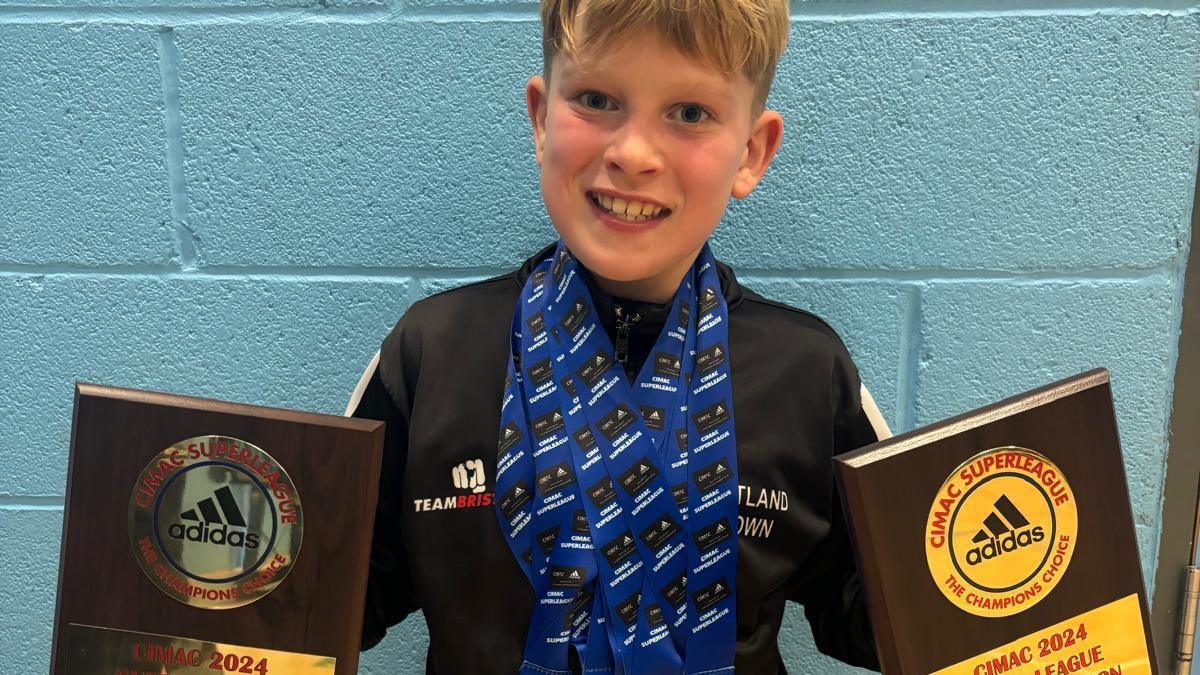 Theo wearing lots of medals around his neck and holding two trophies, one in each hand. He is smiling at the camera