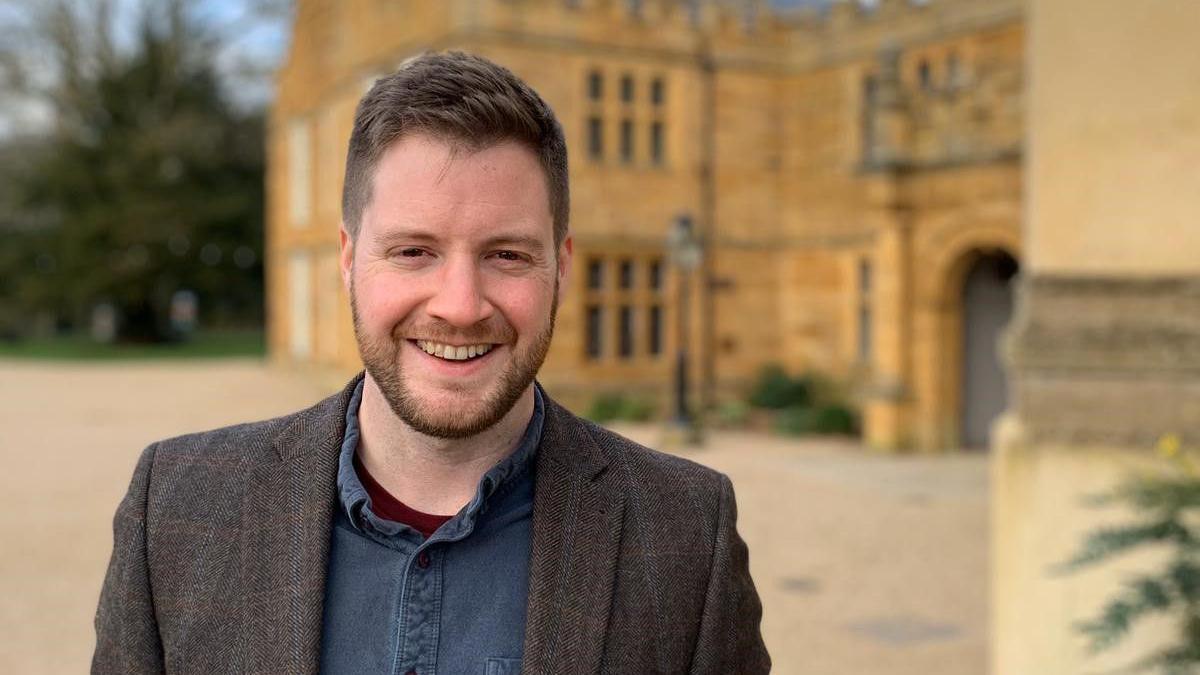 A man looking a the camera a smiling. He is wearing a blue shirt and brown jacket. Behind is a sandstone stately home.
