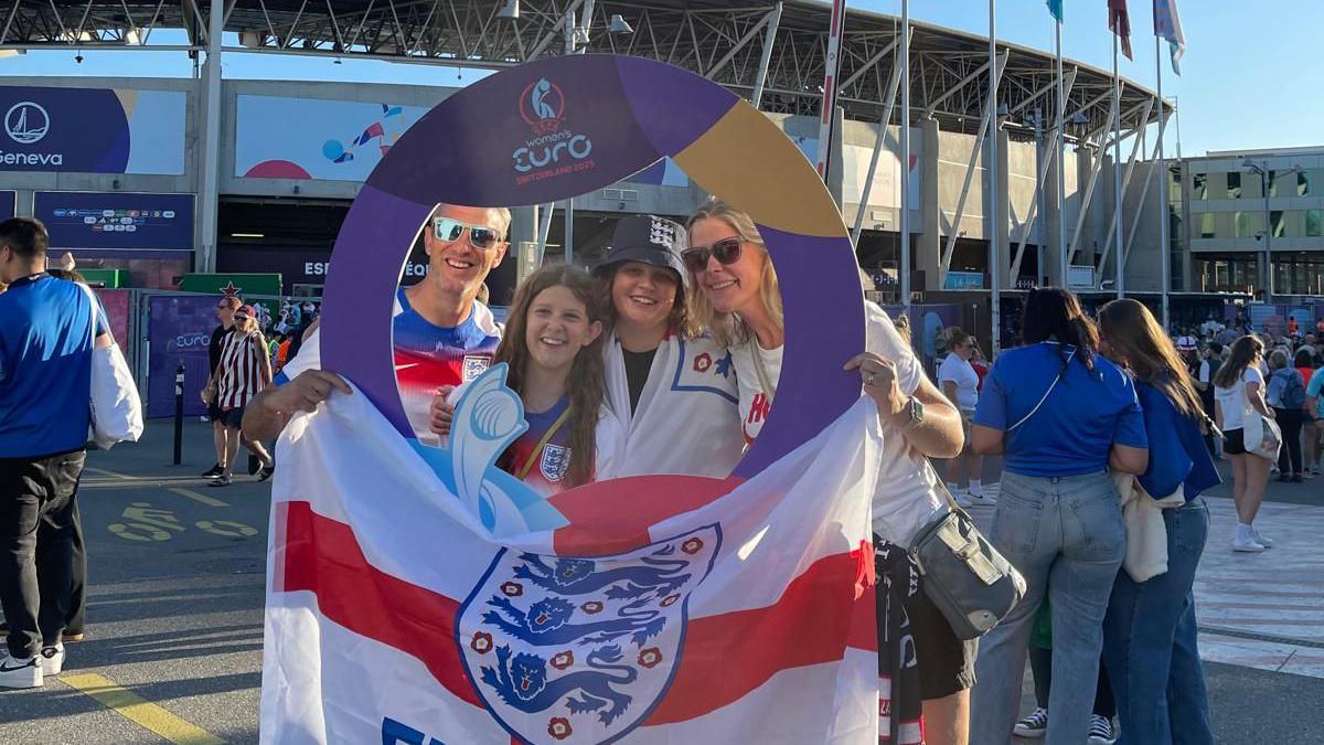 Amy, Arlo, Flo and Neil standing outside a stadium. They're holding up an England flag and posting inside a cutout with the Women's Euros logo on it. It's a sunny day and they're ALL smiling at the camera.