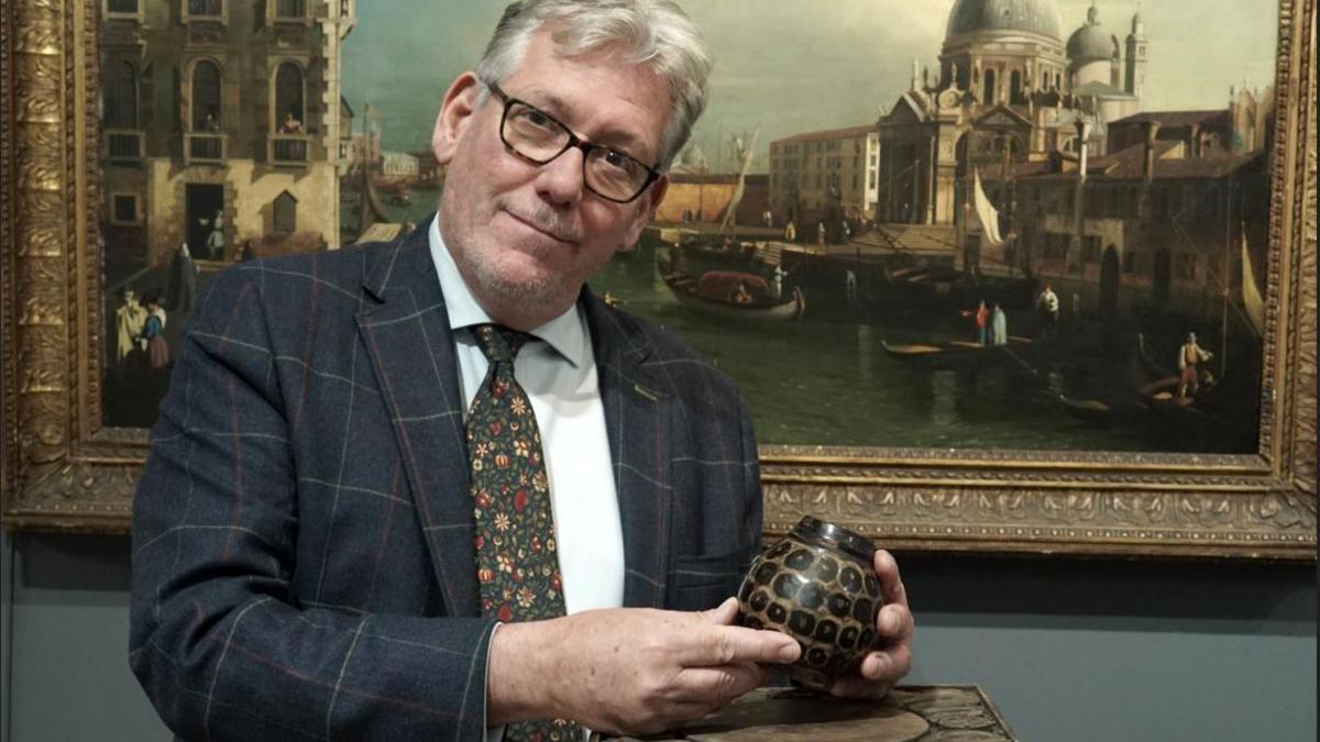 Anderson Garland holding the jar. He is wearing a suit and floral tie. The stone jar fits in its hand. It has a tortoiseshell design and is dark brown in colour. A landscape painting of canals in Venice is behind him.