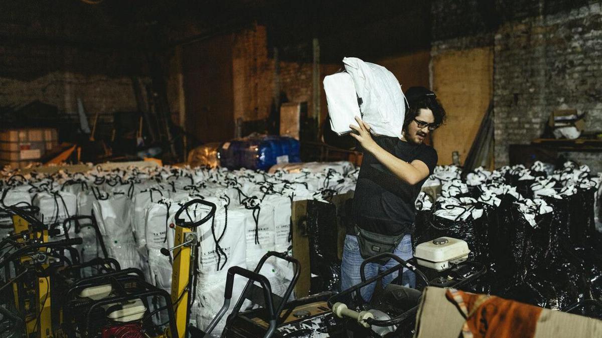 A man in a black t-shirt hoists a white sack over his shoulder. Behind him are rows and rows of sacks and other equipment. He seems to be in a warehouse. 