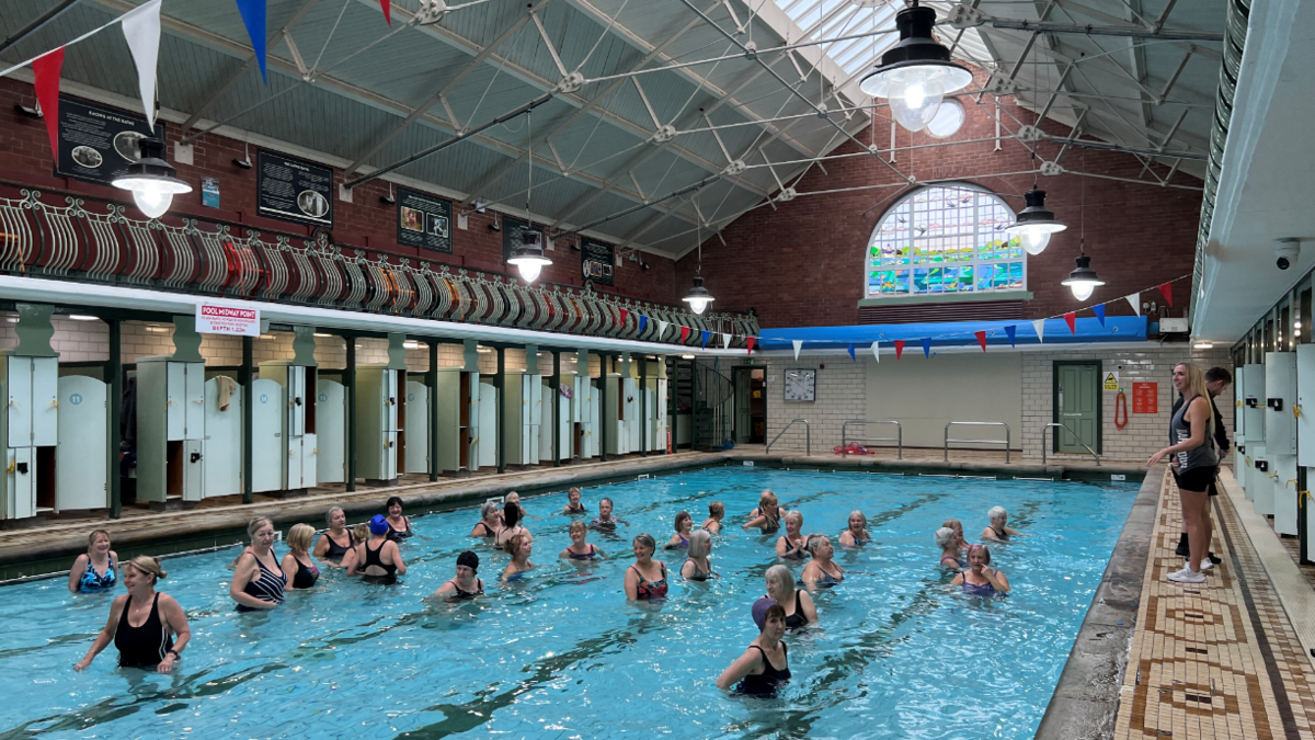 An aqua-aerobics class takes place at Bramley Baths swimming pool in Leeds. Around 30 people are in the water. A member of staff instructs from the tiled sidelines.