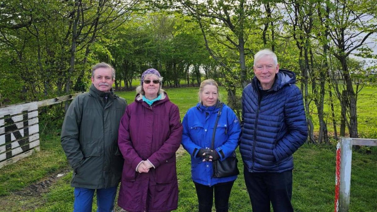 A group of campaigners, two men and two women,  in colourful coats stand smile at the camera on land in front of where the homes are proposed