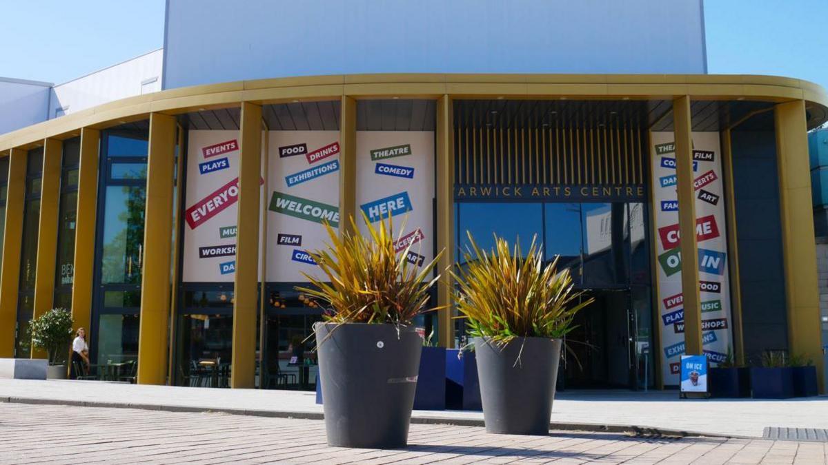 The entrance of Warwick Arts Centre with two large plant pots in the foreground. The name of the centre is written in gold lettering above the door, and the entrance curved, with large golden columns in front and high windows.