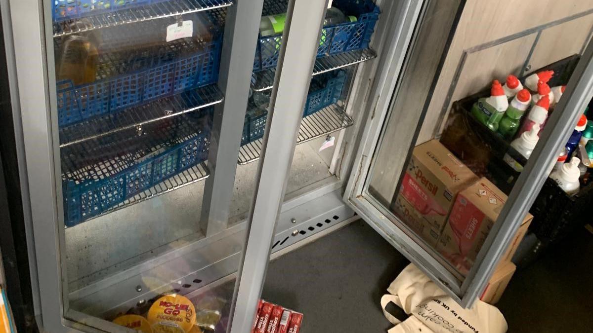 A glass double-doored fridge left open with a tote bag and some food packages on the floor. The fridge shelves are largely empty. There is a box of cleaning supplies to the right which look untouched.