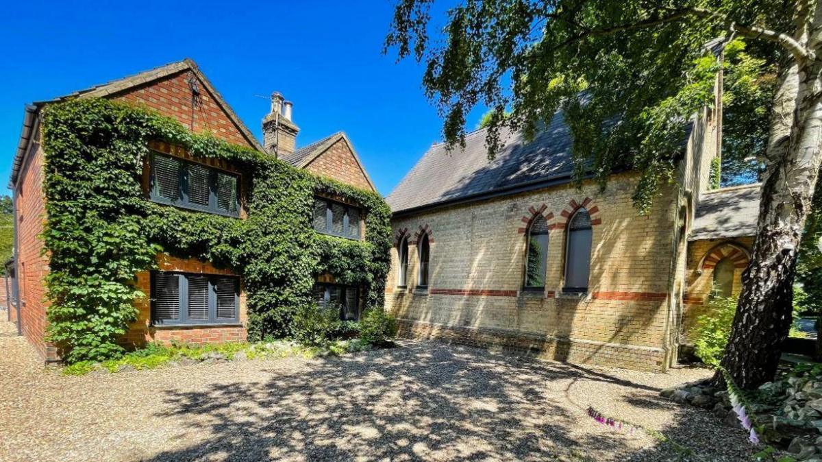 A two-storey brick building covered in thick green ivy next to a lighter-coloured brick building with arched windows. The buildings are separated by a gravel driveway. The sky is blue and the trees are casting shadows on the ground.