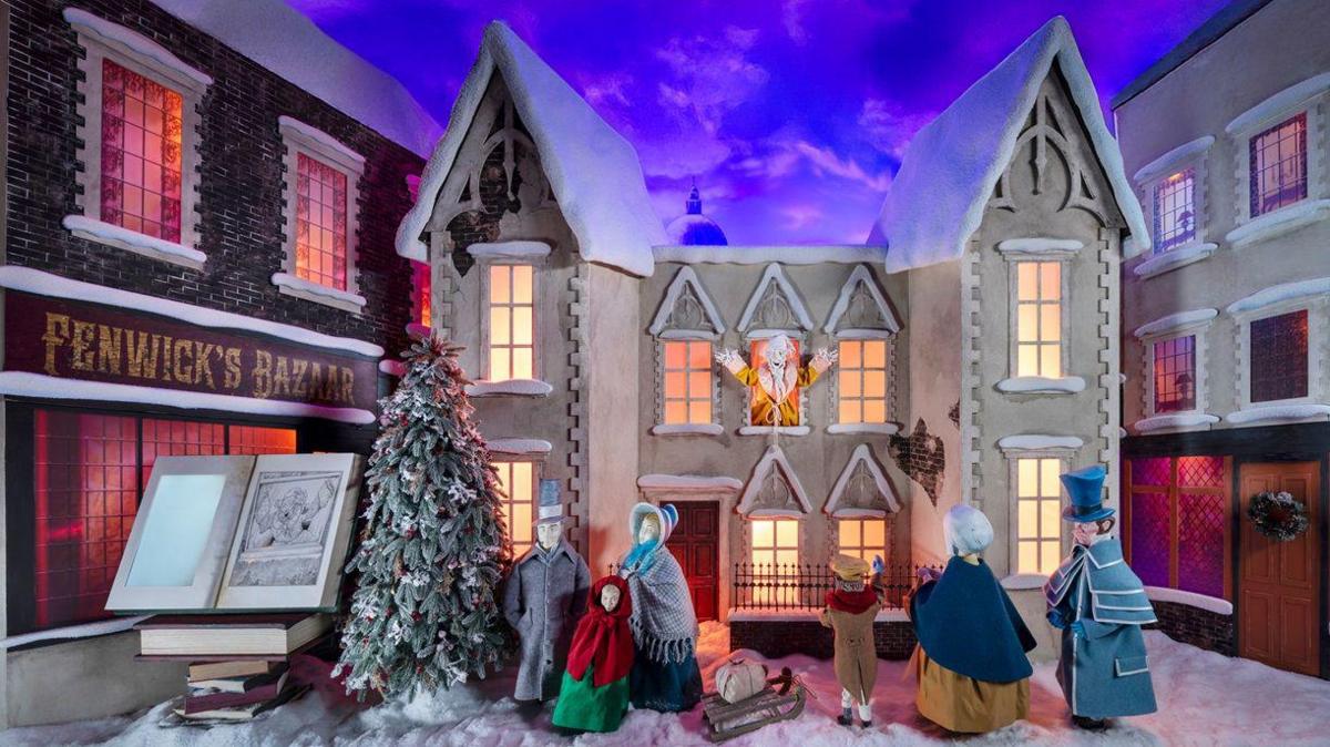 A scene showing figurines outside a house. Scrooge is in the middle window of the house with his arms outstretched. It is snowing and the characters wear Victorian clothing.