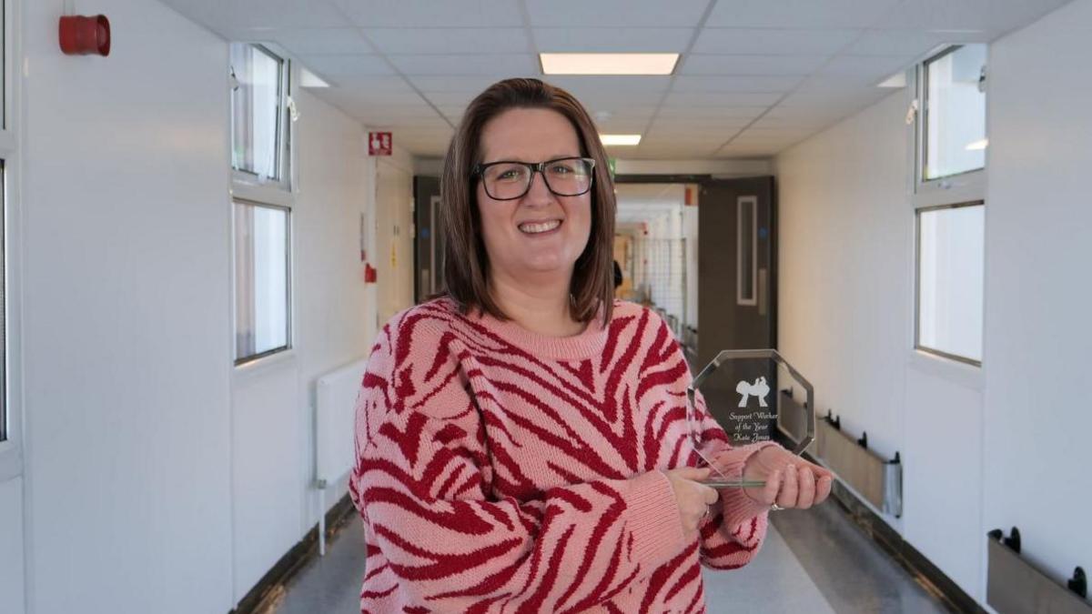 Woman wearing a pink and red animal print jumper holds an award-a sculpture made of glass and she stand in a hospital corridor. 