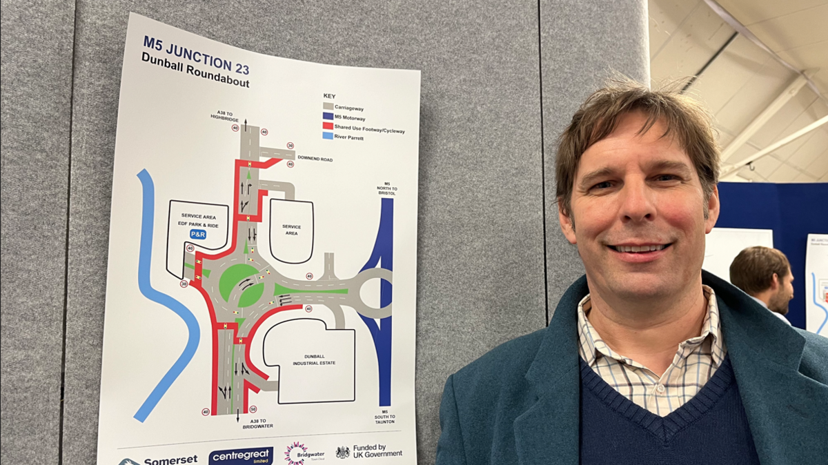 Councillor Richard Wilkins standing next to a poster about the planned works on the roundabout. He is wearing a blue sweater and a blue coat.