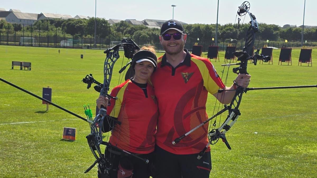 Aalin George and Ethan Moore wearing team Isle of Man red and yellow tops and holding up their archery bows. The archery field with its targets and markers is laid out on grass behind them.