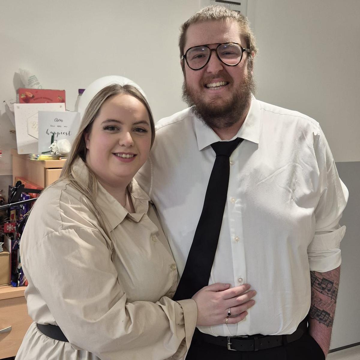 A blond-haired woman stands next to a man with brown hair and a beard. She has her right hand across his stomach showing a wedding band. The man wears a white shirt and black tie. Both smile at the camera