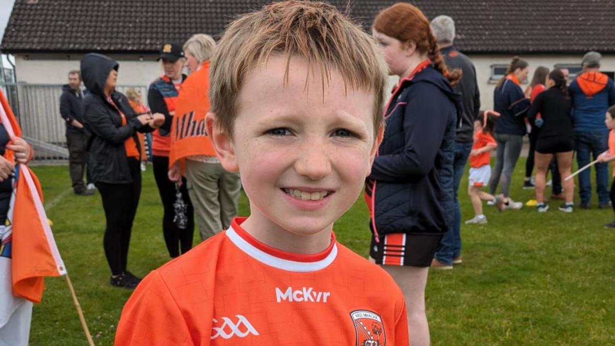 A young boy with blonde hair in an orange Armagh jersey smiles at the camera