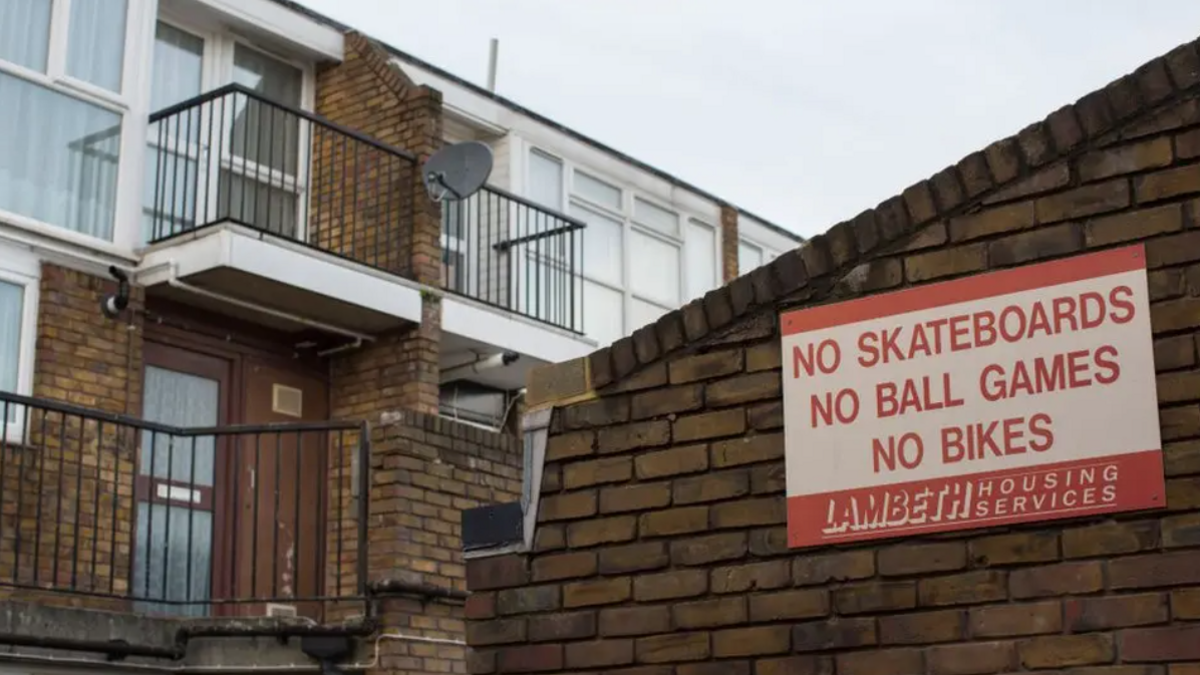 A brick wall with a red and white sign on it, saying "no skateboards, no ball games, no bikes". There's a block of flats behind the sigin.