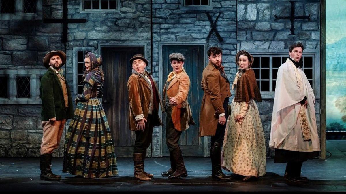 a crowd of men and women in victorian dress singing on the stage while stood in front of a brick house set designed in the background