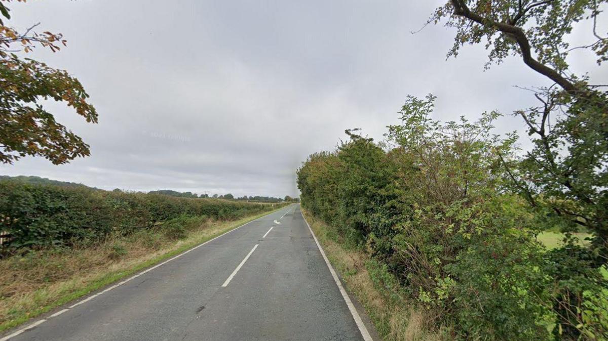 Google street view image of A18 Barton street road. The road is empty and stretches out in a straight line with bushes either side of the road.