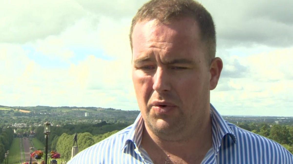 Andrew Keys is a a middle aged man with short light brown hair. He is wearing a blue and white striped shirt with the top two buttons undone. He is standing at the top of a large hill covered in green grass and trees in the background. 