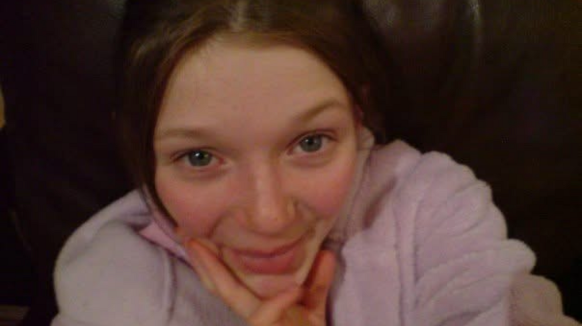 A photograph of a young teenage girl. She is posing for the camera and holding her face, smiling. She wears a lilac coloured jumper.