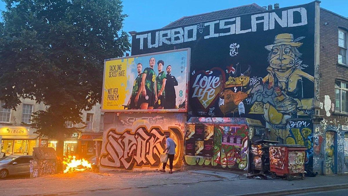 A graffiti wall and advertising board with a small patch of land in front, with a bonfire and a man walking by with a shopping bag.