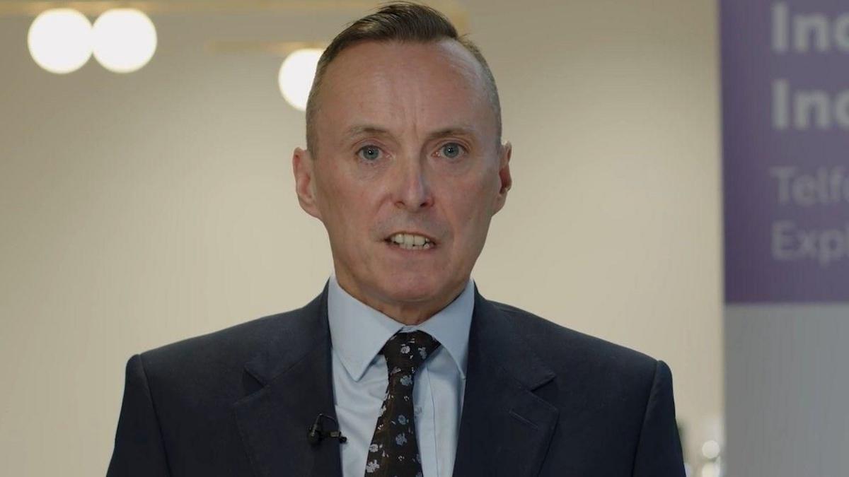 Tom Crowther standing wearing a black suit jacket and black tie with flowers on it. He is also wearing a light blue shirt