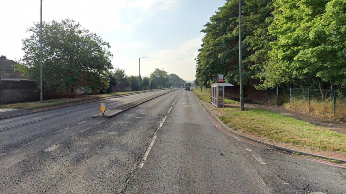 A street view image of a dual carriageway with a T-junction to the right. A bus shelter is also visible on the right with trees and lampposts on both sides of the road. 
