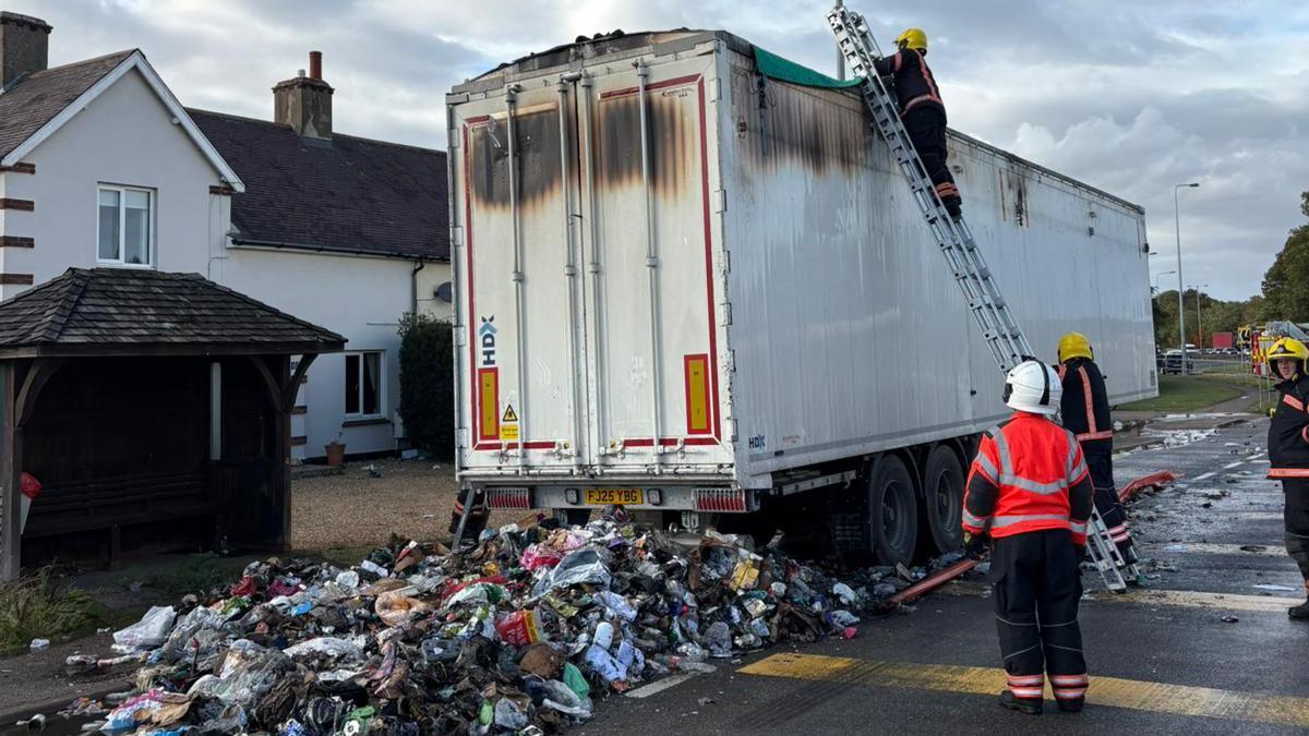 Rubbish on the floor at the back of the lorry. The back doors have been closed and a firefighter is stood at the top of a ladder looking into the lorry from above. The outside of the white lorry has been charred and blackened around the top edges. 