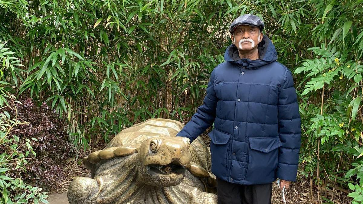 Chacko Abraham Thenakarayil wearing a blue padded coat and cap standing next to a large turtle statue